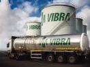 Caminhão tanque da Vibra Energia, com destaque para o logo em verde, e tonéis gigantes ao fundo, alusivo aos resultados do 4º trimestre da companhia