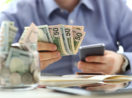 Homem tem um celular na mão e algumas notas de dinheiro, alusivo ao processo de comprar moeda estrangeira