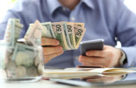 Homem tem um celular na mão e algumas notas de dinheiro, alusivo ao processo de comprar moeda estrangeira