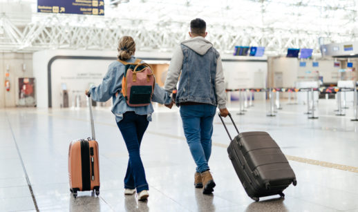 Um homem e uma mulher andam por um aeroporto com suas malas, em alusão a uma viagem de férias