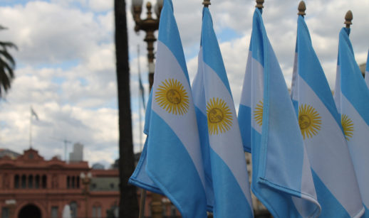 Quatro bandeiras da Argentina enfileiradas em primeiro plano com a Casa Rosada desfocada ao fundo e céu nublado, alusivo ao novo imposto do país