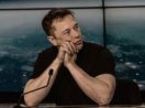 Elon Musk, que quer comprar o Twitter, de camiseta preta e mãos cruzadas, apoiando rosto sobre elas e os cotovelos em mesa perto de microfone