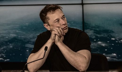 Elon Musk, que quer comprar o Twitter, de camiseta preta e mãos cruzadas, apoiando rosto sobre elas e os cotovelos em mesa perto de microfone