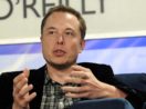 Elon Musk, que cortará salário de direto do Twitter, de terno preto e camisa xadrez cinza por baixo, gesticulando