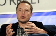 Elon Musk, que cortará salário de direto do Twitter, de terno preto e camisa xadrez cinza por baixo, gesticulando