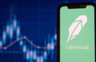 Tela de celular com o logo do app da corretora Robinhood, em primeiro plano, e gráficos de mercado financeiro desfocados ao fundo, alusivo ao mercado de criptomoedas