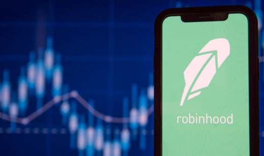 Tela de celular com o logo do app da corretora Robinhood, em primeiro plano, e gráficos de mercado financeiro desfocados ao fundo, alusivo ao mercado de criptomoedas