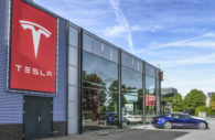 Fachada de concessionária da Tesla, cujas ações foram vendidas por Elon Musk, com destaque para vidros e carro parado na entrada e logo gigante da empresa