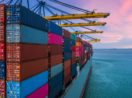 Perspectiva de trás de navio cargueiro lotado com contêineres e pontes rolantes amarelas em cima, com céu rosa e azul, alusivo à balança comercial brasileira em abril