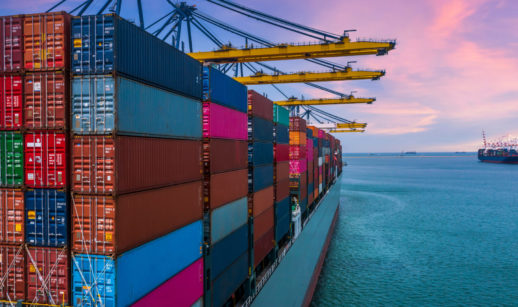 Perspectiva de trás de navio cargueiro lotado com contêineres e pontes rolantes amarelas em cima, com céu rosa e azul, alusivo à balança comercial brasileira em abril