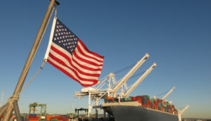 Bandeira dos Estados Unidos em primeiro plano com navio cargueiro ao fundo, alusivo às commodites que ganharam espaço na carteira de BDRs de maio do Safra