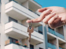 Mão de mulher segurando chave única com o dedo indicador e apartamentos ao fundo, alusivo aos financiamentos imobiliários da Caixa