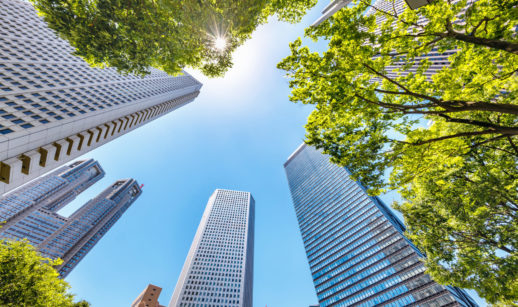 Perspectiva de baixo para cima de prédios espelhados, alusivo ao setor financeiro, com árvores nas bordas, alusivo às ações ESG