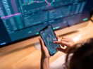 Mãos mexendo no celular em tela do mercado financeiro em frente ao desktop, alusivo à carteira recomendada de ações para investir em junho