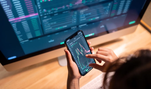Mãos mexendo no celular em tela do mercado financeiro em frente ao desktop, alusivo à carteira recomendada de ações para investir em junho