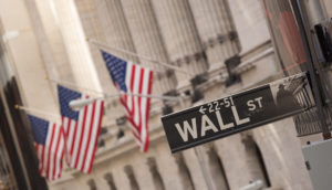 Placa em primeiro plano de Wall Street com bandeiras dos EUA ao fundo em frente a uma das bolsas americanas