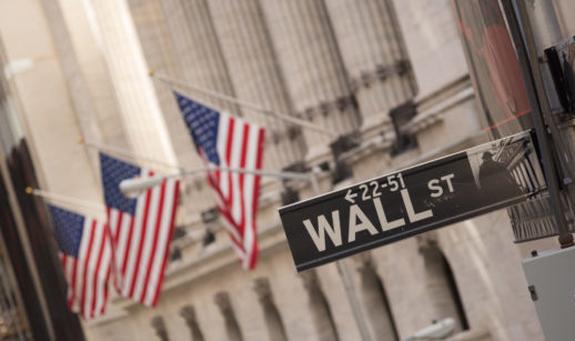 Placa em primeiro plano de Wall Street com bandeiras dos EUA ao fundo em frente a uma das bolsas americanas