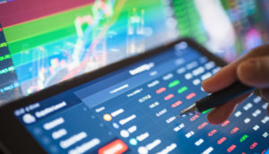 Detalhe de dedo com caneta touch apontando para tela de tablet com PC atrás e gráficos de mercado financeiro, alusivo às recomendações para investir em renda variável