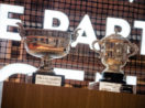 Troféus de Roland Garros, prateados, lado a lado, com fundo simulando rede de tênis, alusivo à premiação