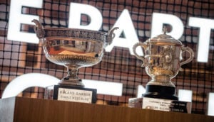Troféus de Roland Garros, prateados, lado a lado, com fundo simulando rede de tênis, alusivo à premiação