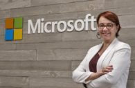 Tânia Cosentino, presidente da Microsoft, à direita da foto, de blazer branco e blusa roxa por baixo, óculos de armação vermelha, sorrindo e logo da Microsoft ao centro