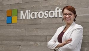 Tânia Cosentino, presidente da Microsoft, à direita da foto, de blazer branco e blusa roxa por baixo, óculos de armação vermelha, sorrindo e logo da Microsoft ao centro