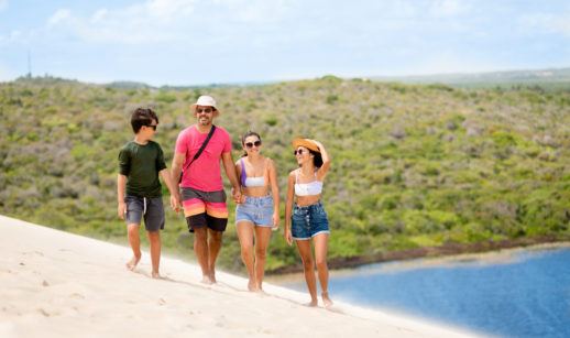Quatro pessoas de mesma família andando sobre dunas de areia com mata atrás, alusivo ao turismo