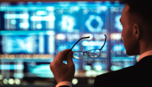 Homem de costas de terno preto e óculos em mãos, olhando para tela de informações do mercado financeiro, alusivo à mudança do cenário fiscal