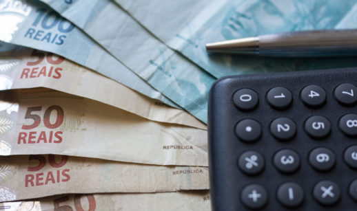 Cédulas de 50 e 100 reais sobrepostas ao lado de caneta e calculadora, alusivo ao investimento no Tesouro Direto IPCA+, atrelado à inflação