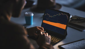 Perspectiva de trás para frente de homem em laptop digitando e vendo gráfico de ações, alusivo às top picks para investir em renda variável