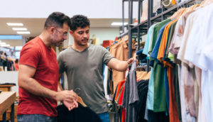 Dois homens de camisa cinza e vermelha mexendo em arara de lojas de roupas, alusivo às vendas nos shoppings da Multiplan
