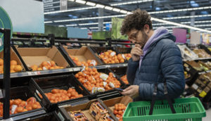 Homem de óculos e jaqueta segurando celular e cesta vazia de supermercado, alusivo à inflação do Reino Unido