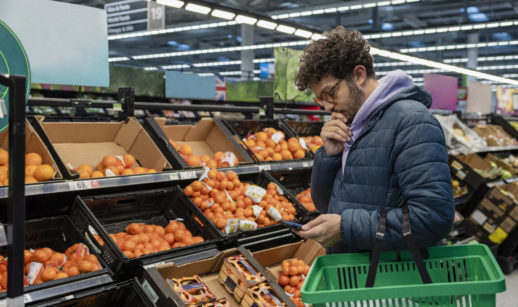 Homem de óculos e jaqueta segurando celular e cesta vazia de supermercado, alusivo à inflação do Reino Unido