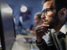 Homem de barba e óculos, de perfil, com mão no queixo, olhando para o computador, alusivo ao escritório de investimentos