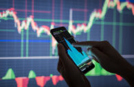 Detalhe de mãos mexendo em celular com gráficos de mercado financeiro e telão atrás, alusivo às ações de tecnologia