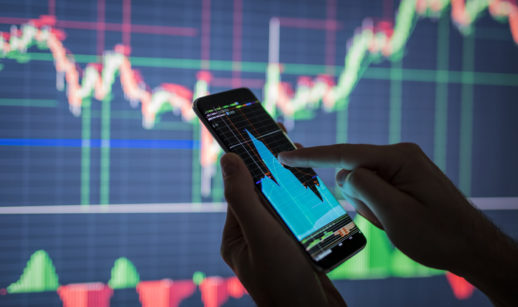 Detalhe de mãos mexendo em celular com gráficos de mercado financeiro e telão atrás, alusivo às ações de tecnologia