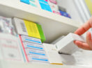 Close de mão pegando medicamento em estante de farmácia, alusivo ao setor farmacêutico