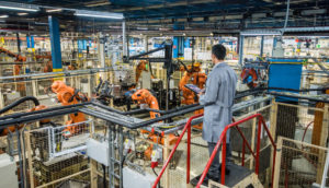 Indústria de máquinas com homem de costas de macacão monitorando com tablet na mão, alusivo à produção industrial de maio