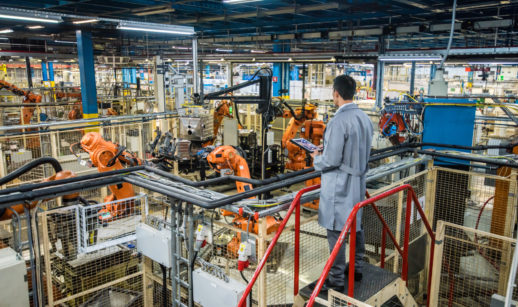 Indústria de máquinas com homem de costas de macacão monitorando com tablet na mão, alusivo à produção industrial de maio