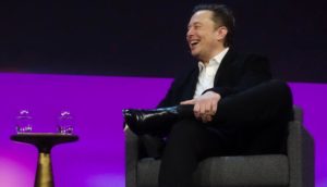 À direita da foto, Elon Musk, sentado, de terno preto com camisa branca sem gravata, olhando para a esquerda e rindo, alusivo ao deboche sobre a desistência de compra das ações do Twitter