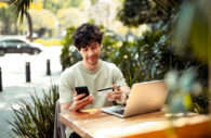 Homem sentado em mesa de madeira com celular, cartão de crédito em mãos e laptop em frente, alusivo às transações bancárias por canais digitais