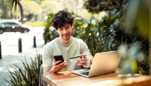 Homem sentado em mesa de madeira com celular, cartão de crédito em mãos e laptop em frente, alusivo às transações bancárias por canais digitais