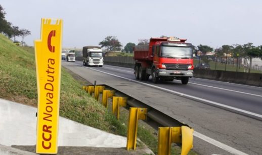 Caminhão vermelho à esquerda andando pela rodovia Presidente Dutra, com destaque para totem amarelo à esquerda com o logo da CCR, que teve fatia comprada por Itaúsa e Votorantim
