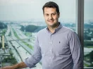Marcelo França, CEO da Celcoin, de camisa listrada e mão apoiando em janela, sorrindo