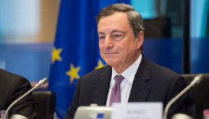 Mario Draghi, de terno preto e gravata roxa, sorrindo de boca fechada e bandeira da União Europeia em segundo plano