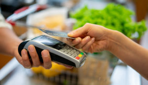 Close de mão usando o cartão de crédito em máquina com verduras ao fundo, alusivo à expansão do crédito no Brasil