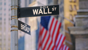 Placa de Wall Street, em Nova York, nos EUA. com bandeira do país ao fundo, alusivo à diversificação internacional em agosto com a Carteira Global