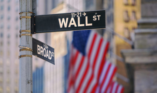 Placa de Wall Street, em Nova York, nos EUA. com bandeira do país ao fundo, alusivo à diversificação internacional em agosto com a Carteira Global