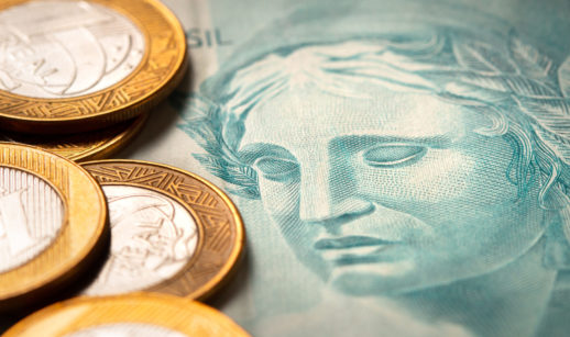 Close de nota de 100 reais em azul com moedas de 1 real do lado esquerdo, alusivo ao investimentos nos títulos do Tesouro Direto