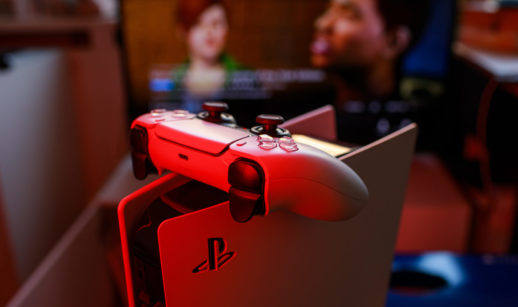 Detalhe de PlayStation 5 com controle em cima iluminados de vermelho e TV ao fundo, alusivo à subida de preço realizada pela Sony